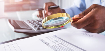 Businessman Holding Magnifying Glass Over Invoice Close-up Of A Businessman Holding Magnifying Glass Over Invoice Using Calculator
